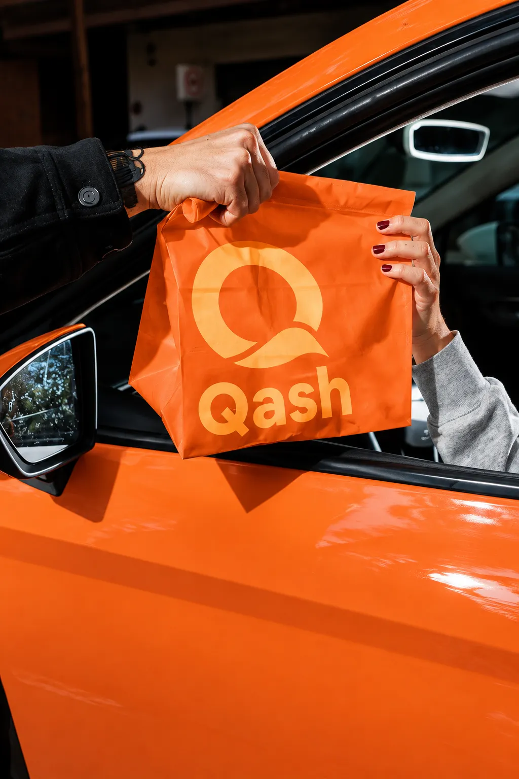 Tas kertas oranye bertuliskan Qash diserahkan ke jendela mobil oranye