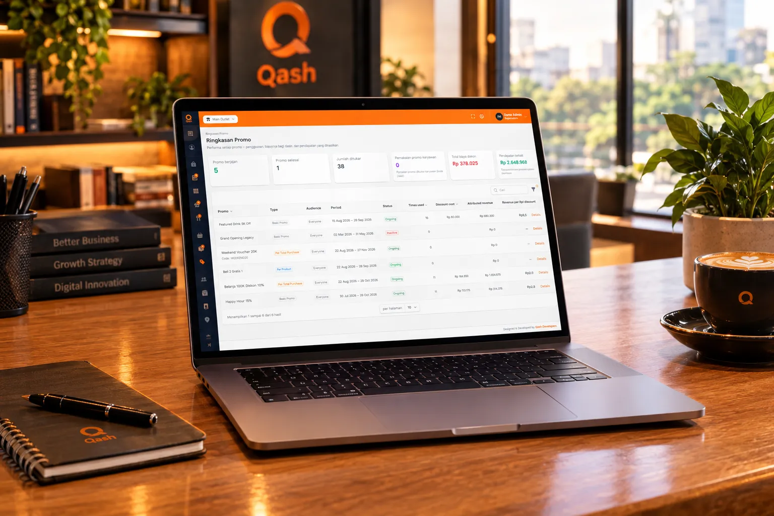 Laptop di meja kerja menampilkan ringkasan promo Qash, di samping buku dan kopi